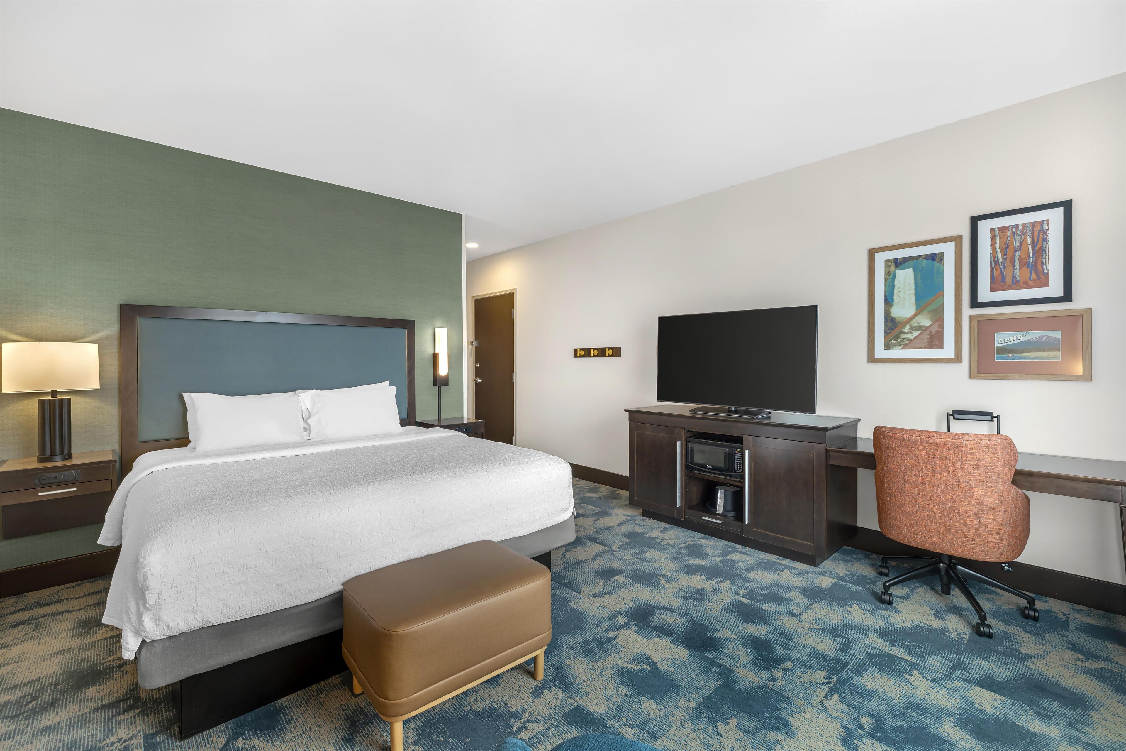 A hotel room with a bed, a desk with a television, a chair, and artwork on the wall.