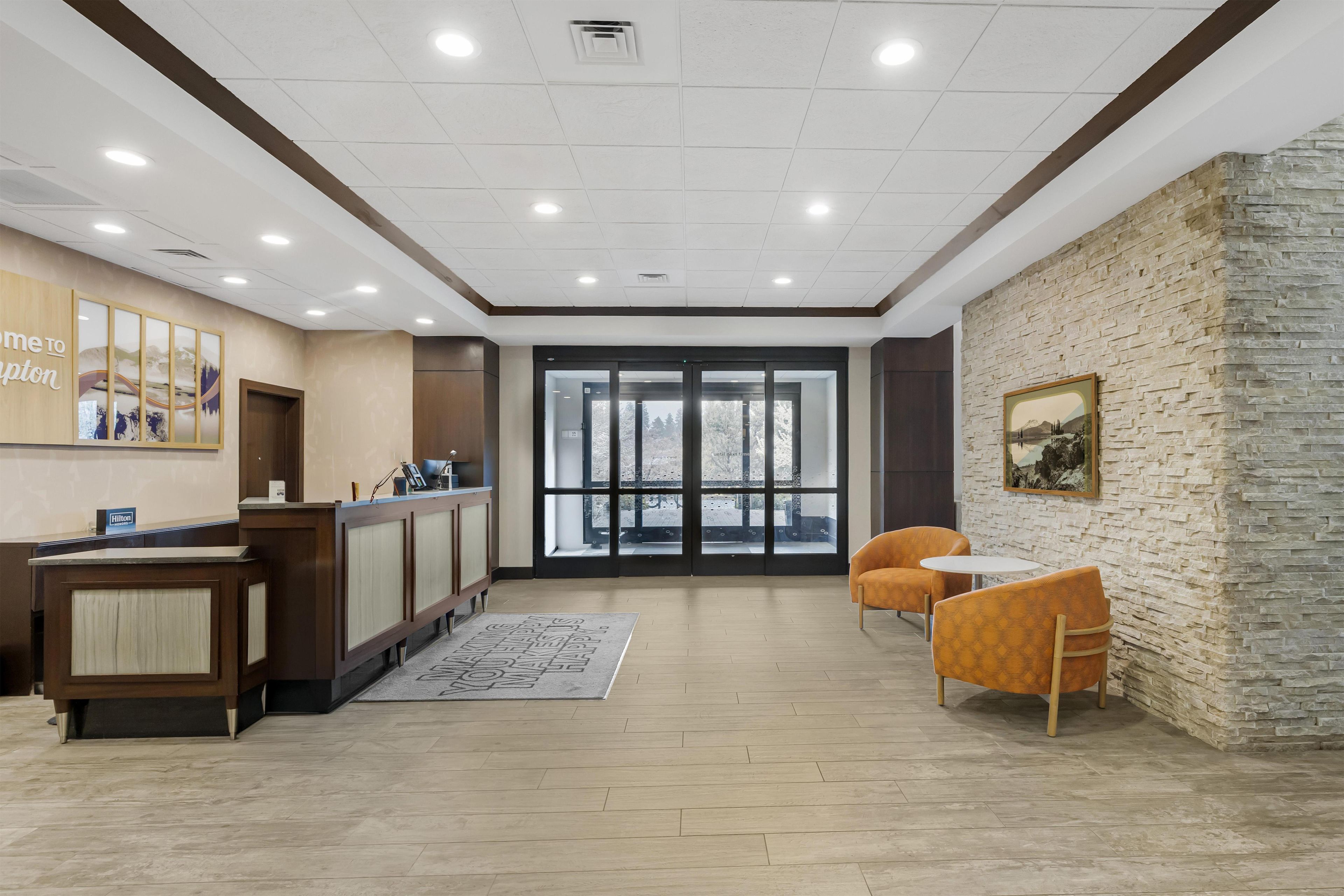 A hotel reception area with a wooden reception desk, a seating area with two chairs, a large window with a view, and a stone wall with a framed picture.