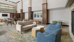 A spacious lobby with a sofa, chairs, a coffee table, a fireplace, a large painting, and a bar area. - Hampton Inn & Suites Bend (Bend)