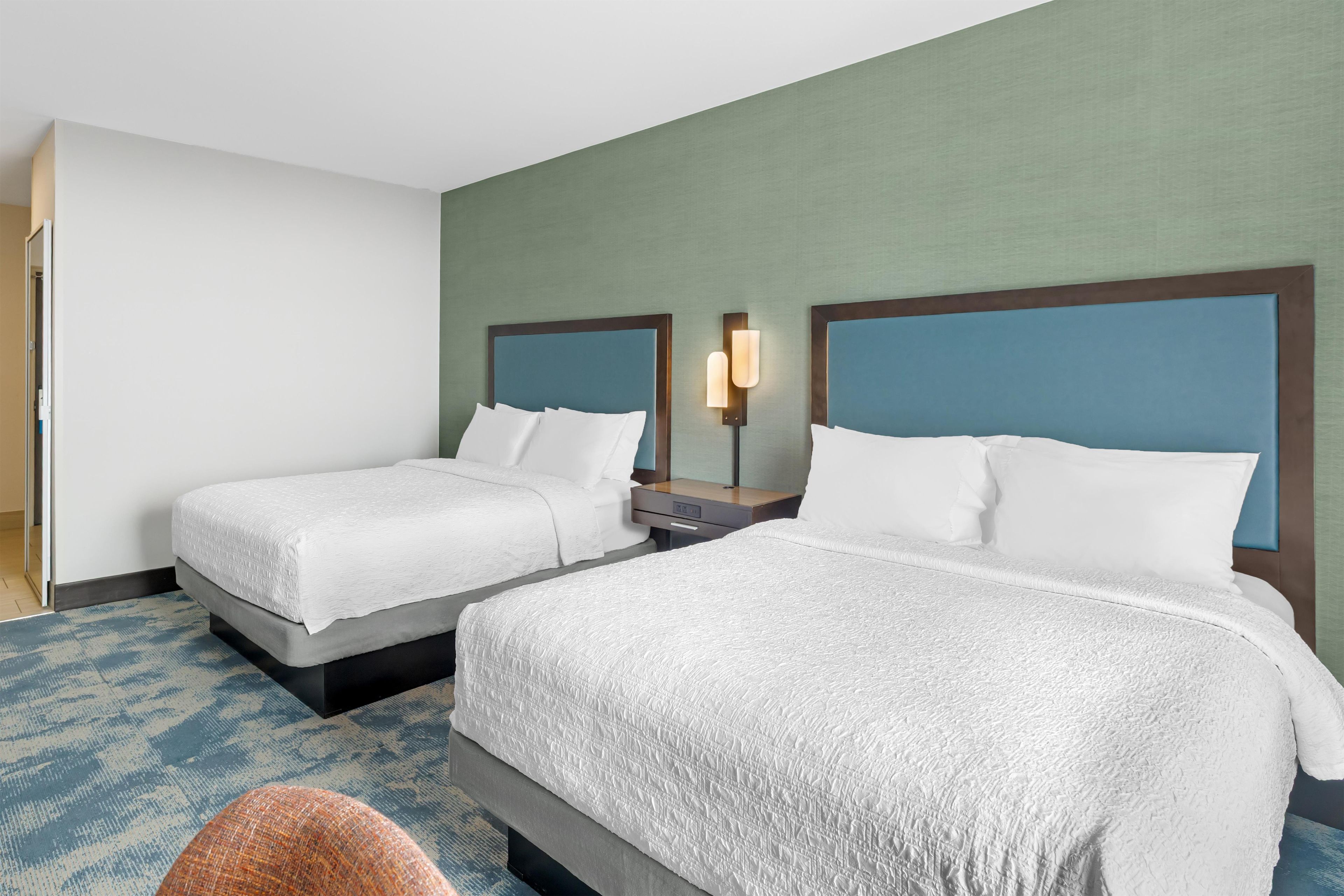 Quarto de hotel com duas camas, uma mesa de cabeceira e uma luminária fixada na parede.