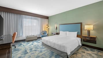 A hotel room with a bed, a desk, a chair, a window with curtains, a wall-mounted lamp, and a bedside table with a lamp.