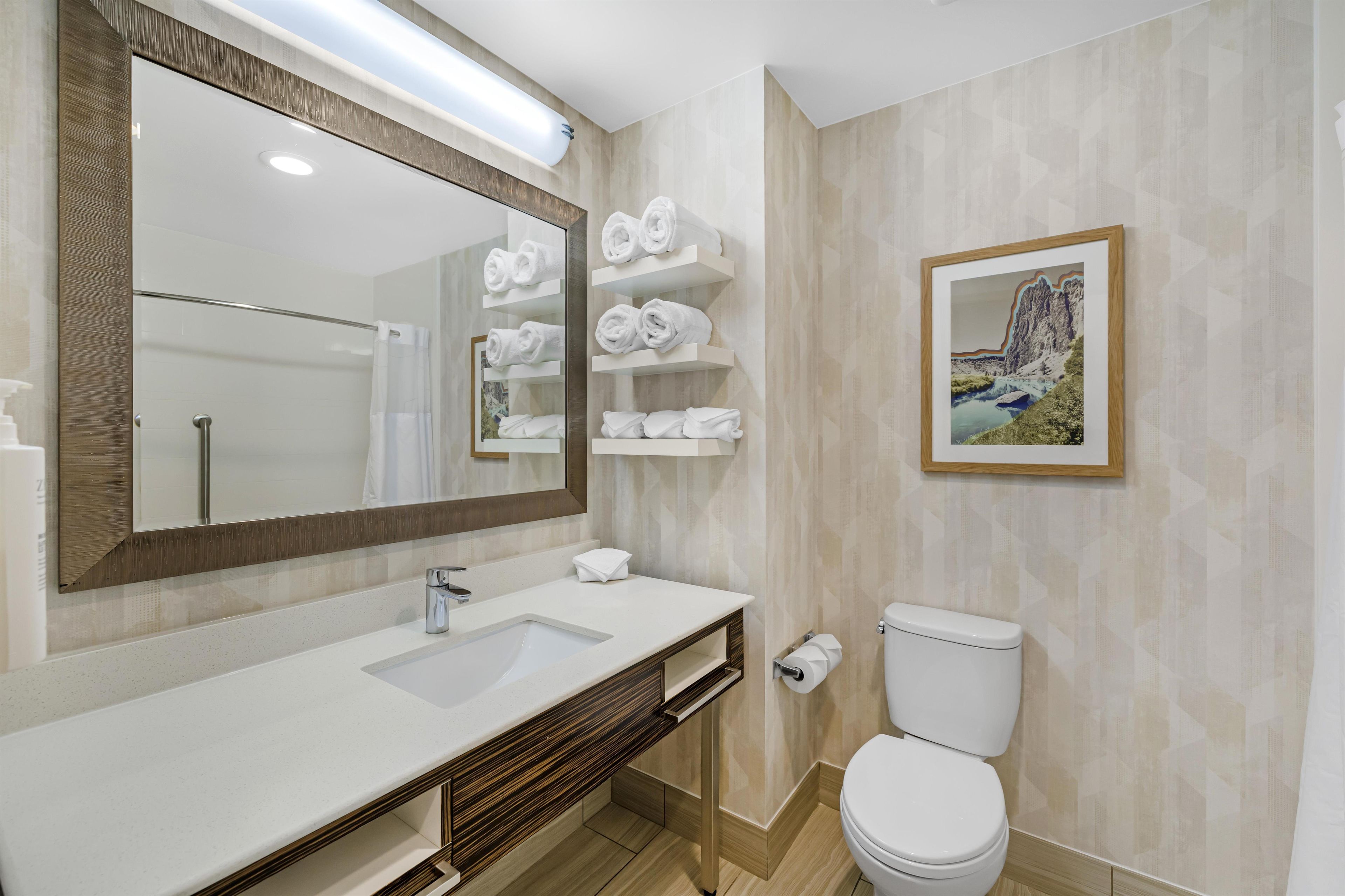 A bathroom with a large mirror, white sink, wooden vanity, towel racks, toilet, and a framed picture on the wall.