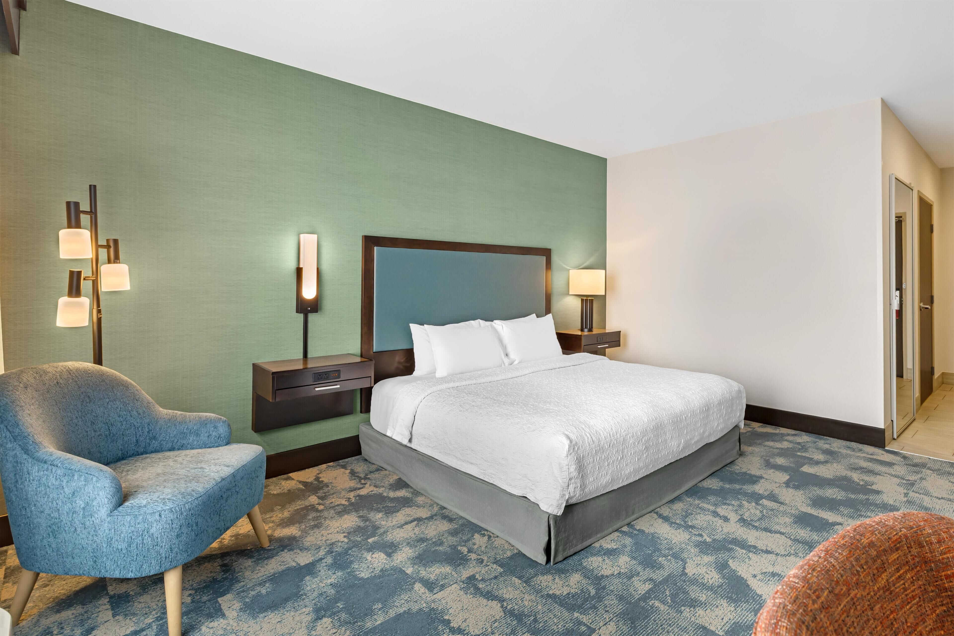 A hotel room with a bed, a chair, a nightstand, and a lamp.