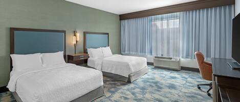 Habitación de hotel con dos camas, un escritorio, una silla y un ventanal con cortinas.