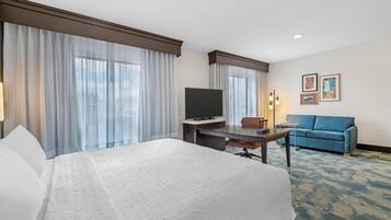 Quarto de hotel com cama, escrivaninha, televisão, sofá e janelas amplas com cortinas.