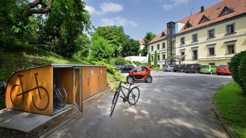 A bicycle storage area with a bike inside, a small building with a bicycle symbol, and a parked car in front of a building with the sign "HOTEL MAXMILIAN".