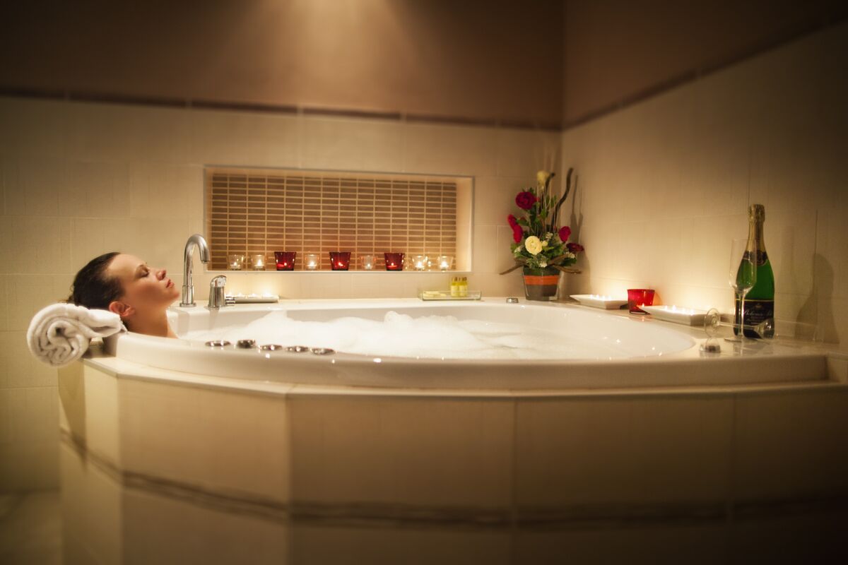 A person relaxing in a large bathtub with a window above and candles on the windowsill.
