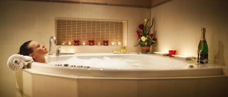 A person relaxing in a large bathtub with a window above and candles on the windowsill.