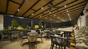 An outdoor dining area with wooden tables and chairs, a ceiling fan, and a view of the surrounding landscape. - Maximilian Lifestyle Resort (Loucen)