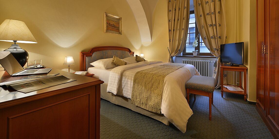 A hotel room with a bed, a desk with a lamp, a television, and a window with curtains.