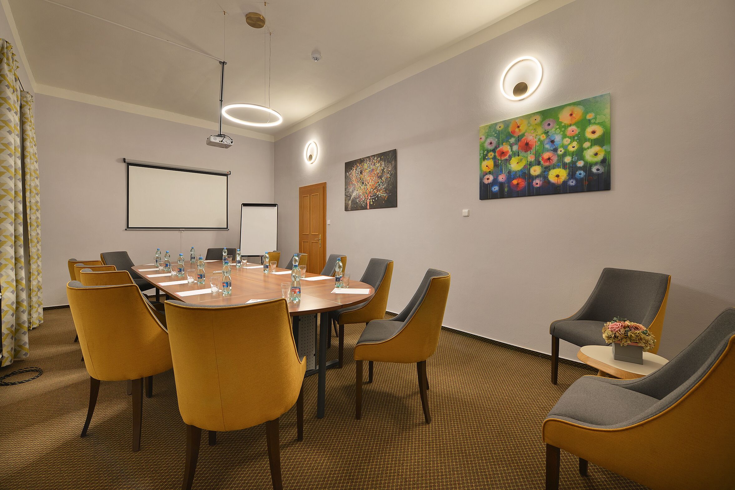 Una sala conferenze con un lungo tavolo, sedie gialle, uno schermo per proiettore e un dipinto floreale al muro.