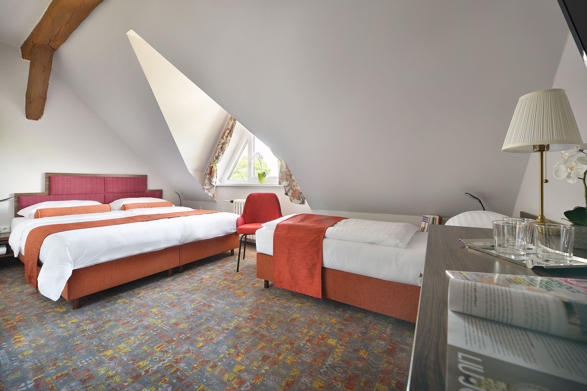 A hotel room with two beds, a red chair, a desk with a lamp, and a window with curtains.