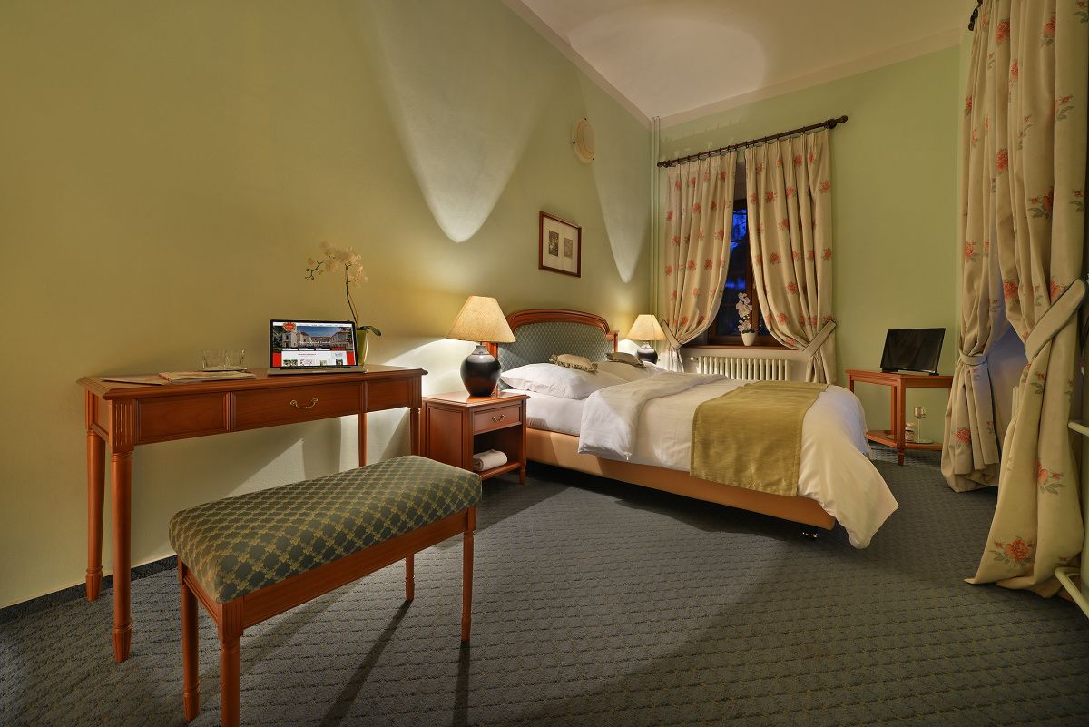 A hotel room with a bed, a desk, a chair, a small table, a lamp, and a window with curtains.