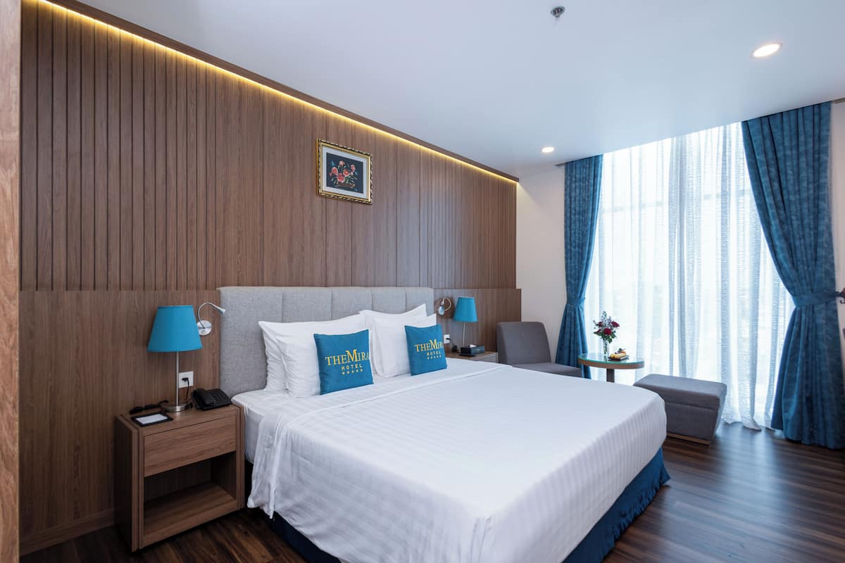 Une chambre d’hôtel avec un grand lit, deux tables de chevet, une petite table d’appoint, une petite table, une chaise et une fenêtre avec des rideaux.