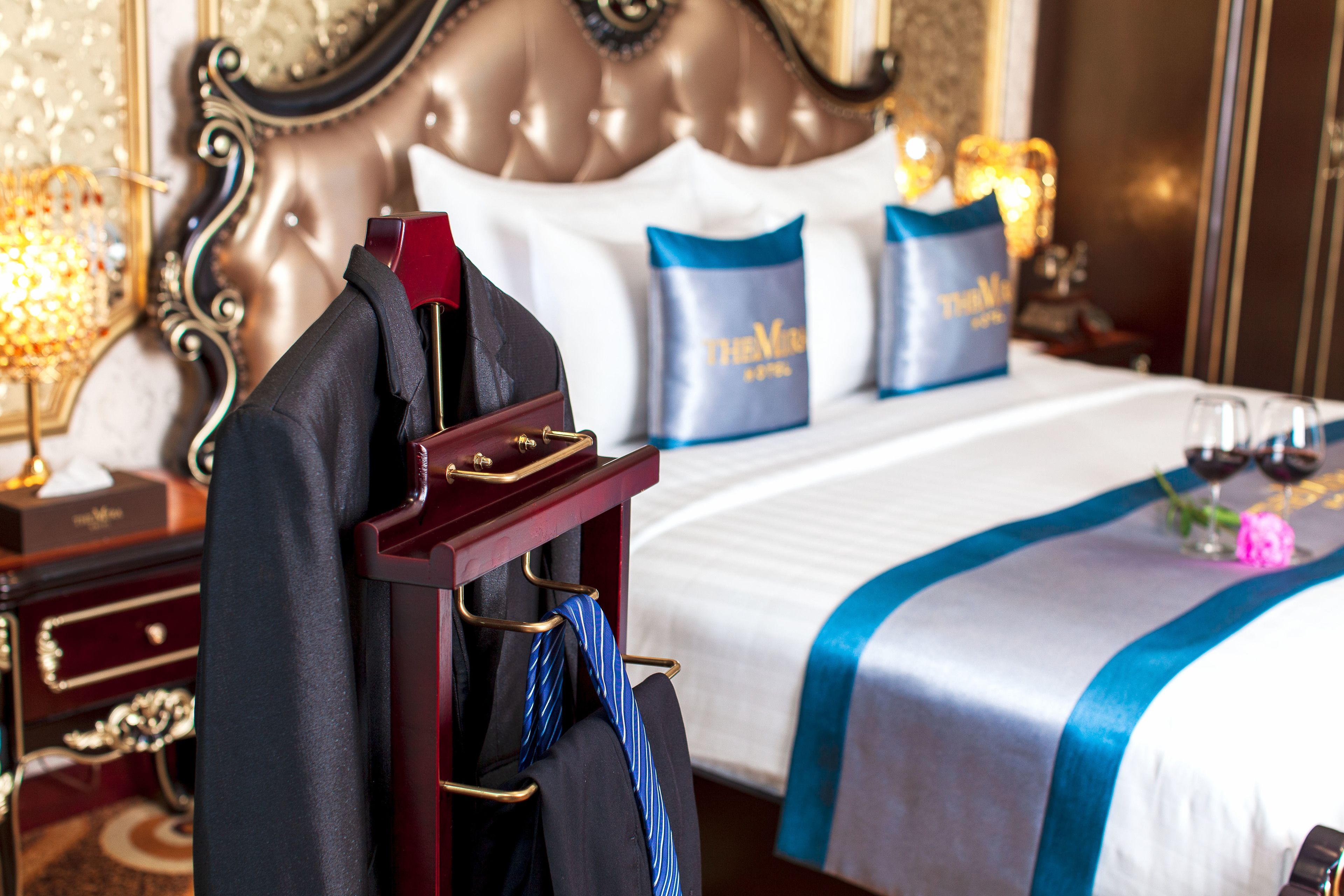 A hotel room with a neatly made bed, a side table with a lamp, and a coat rack with a suit and tie.