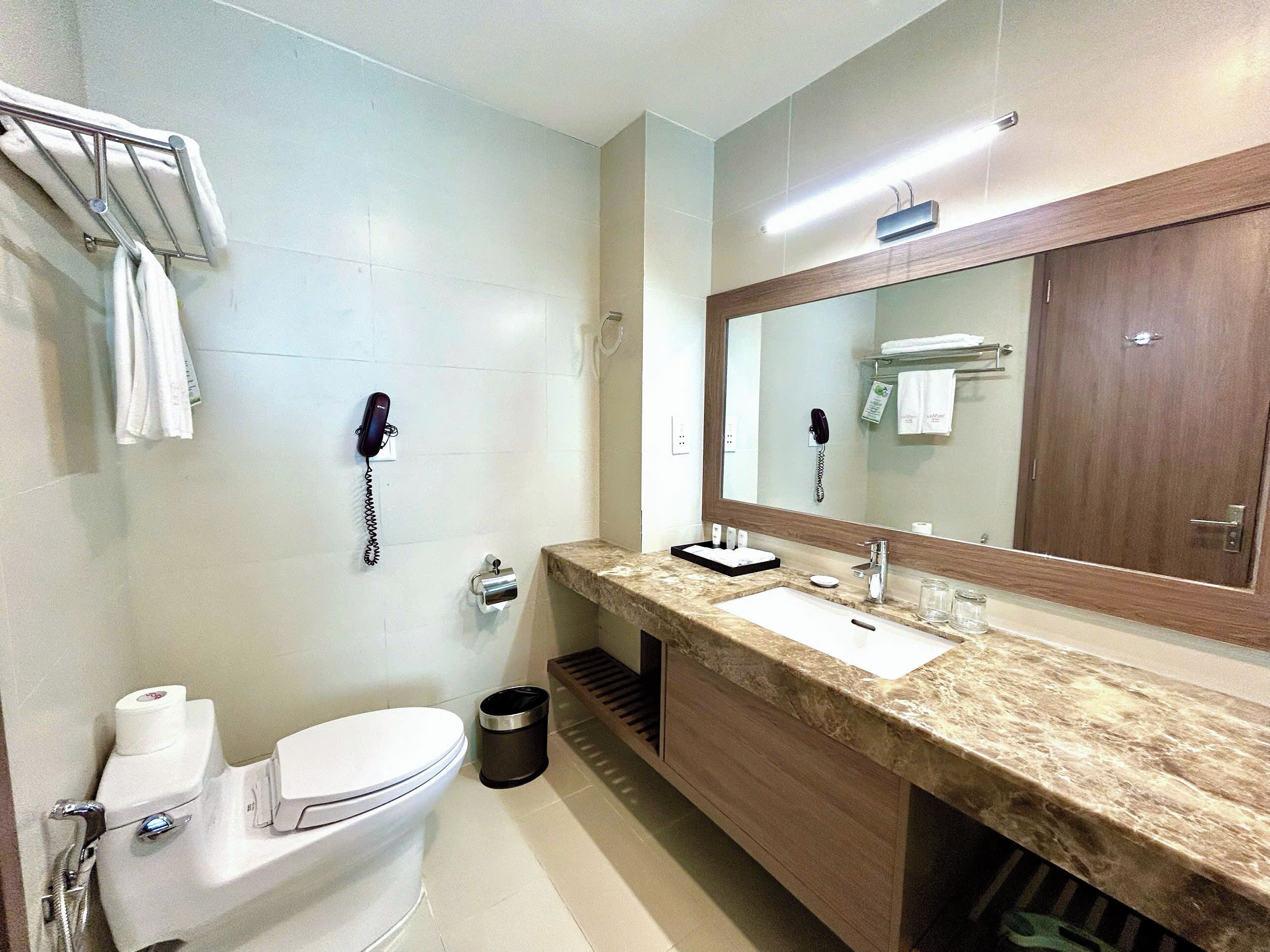 A modern bathroom with a toilet, sink, and mirror. There are two phones mounted on the wall, a trash can, and a towel rack.