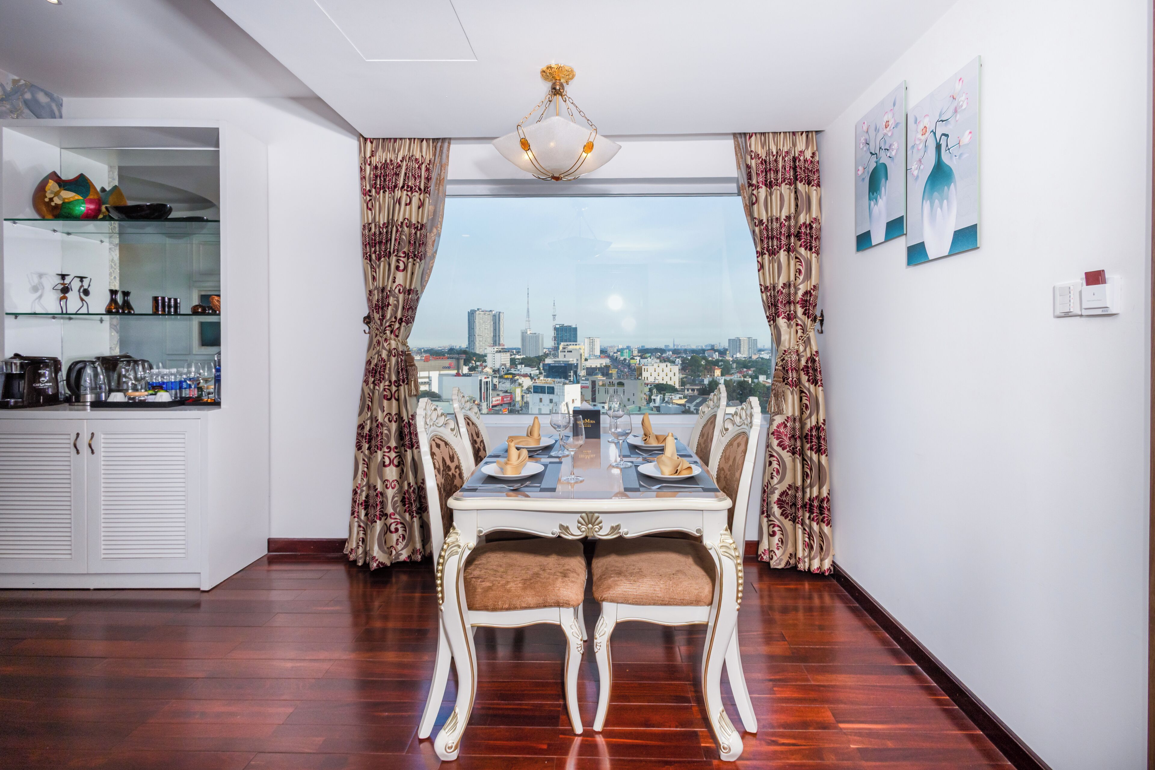 A dining area with a table set for four, a view of the cityscape through large windows, and a kitchen area with cabinets and shelves.