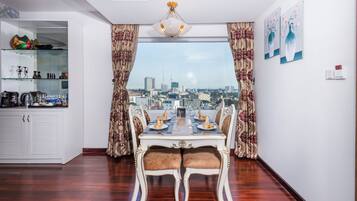 A dining area with a table set for four, a view of the cityscape through large windows, and a kitchen area with cabinets and shelves.