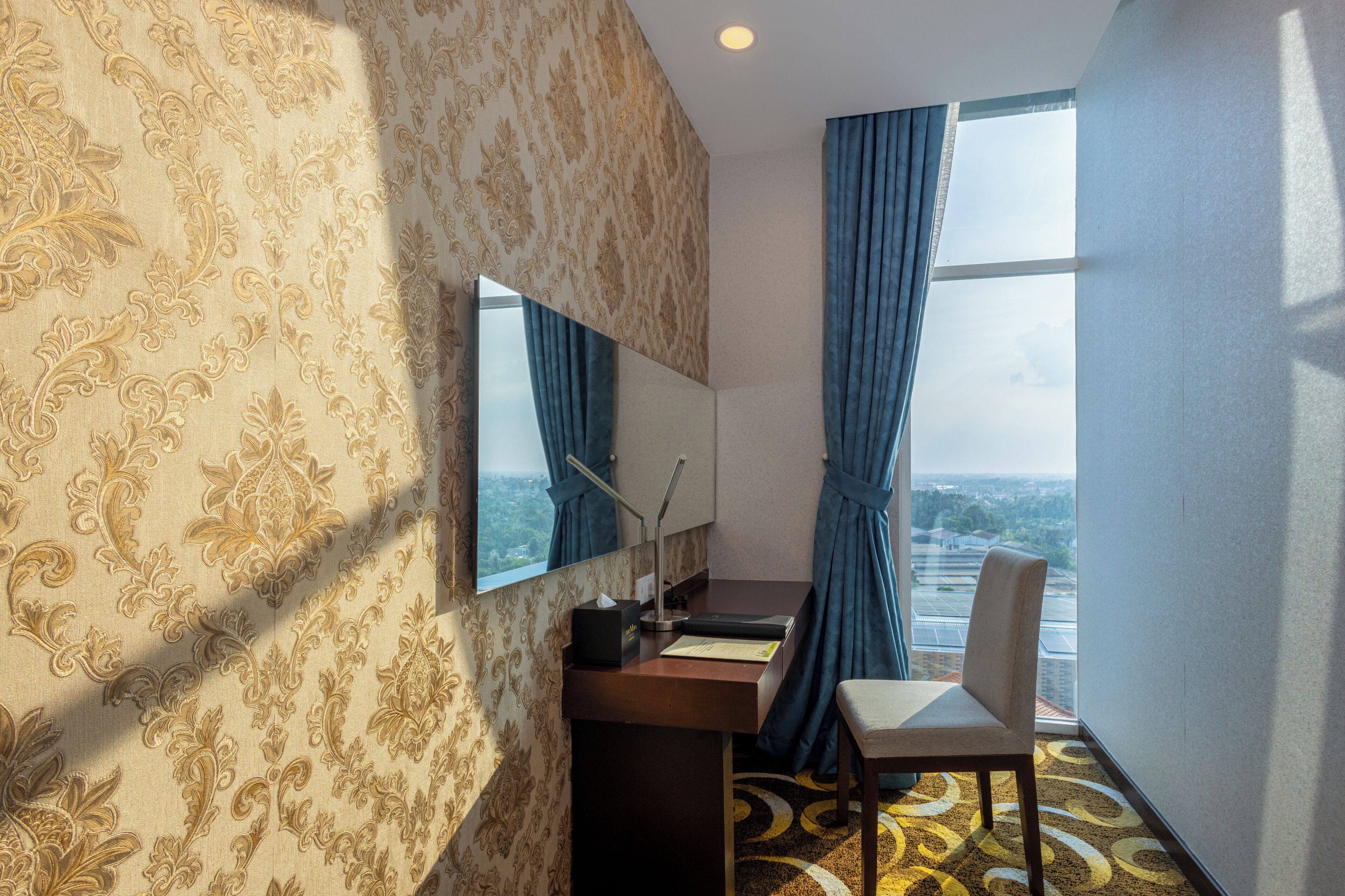 A hotel room with a large window, a desk with a chair, a mirror, and a patterned carpet.