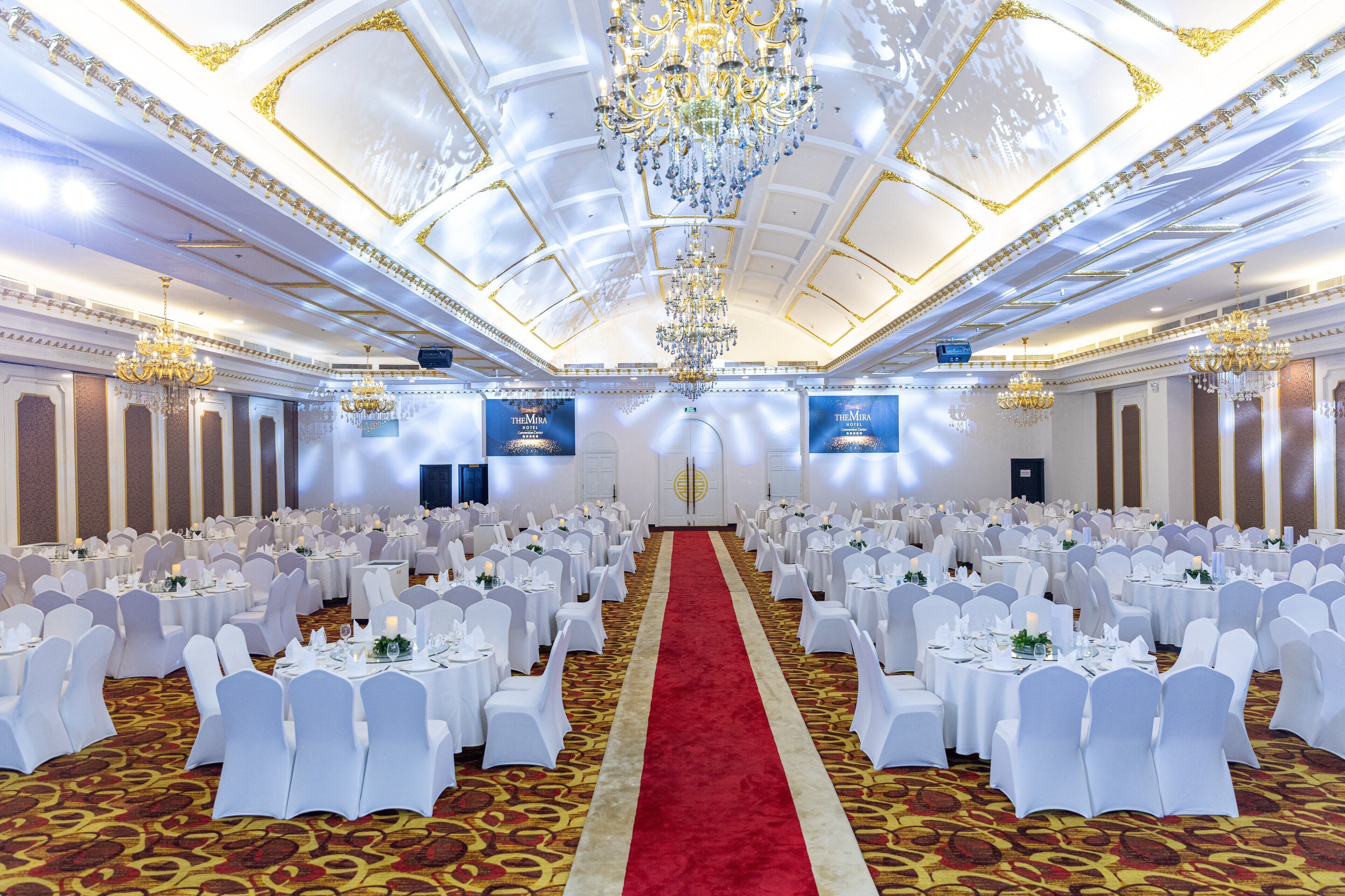 A banquet hall with round tables, white chairs, a red carpet, and chandeliers.