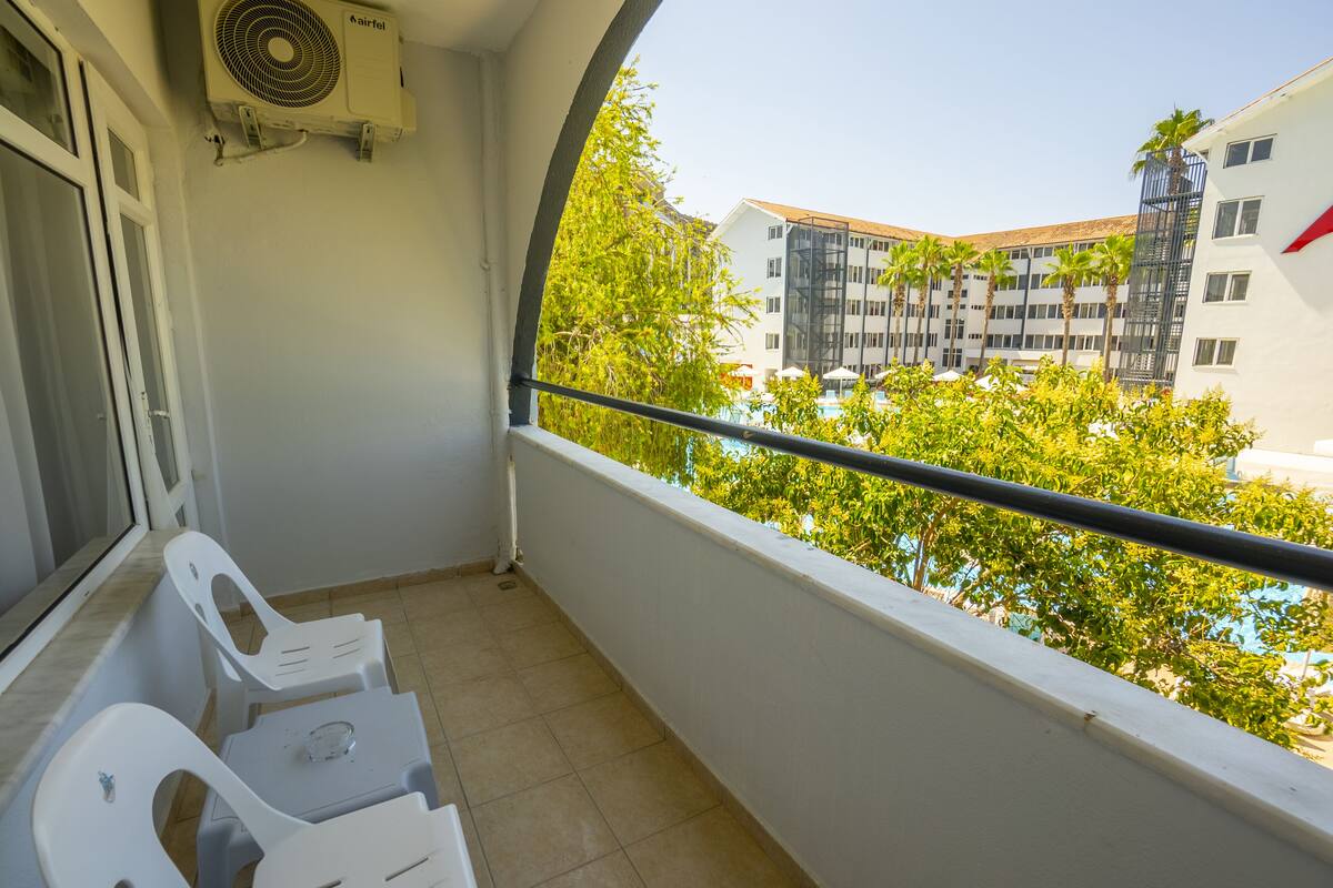 A balcony with white plastic chairs, a small table, and a view of a building with palm trees and a pool.