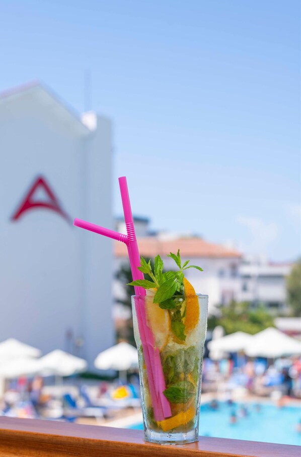 A glass with a pink straw and mint leaves, placed on a wooden surface with a pool and umbrellas in the background. - Armas Bella Sun - All Inclusive (Manavgat)