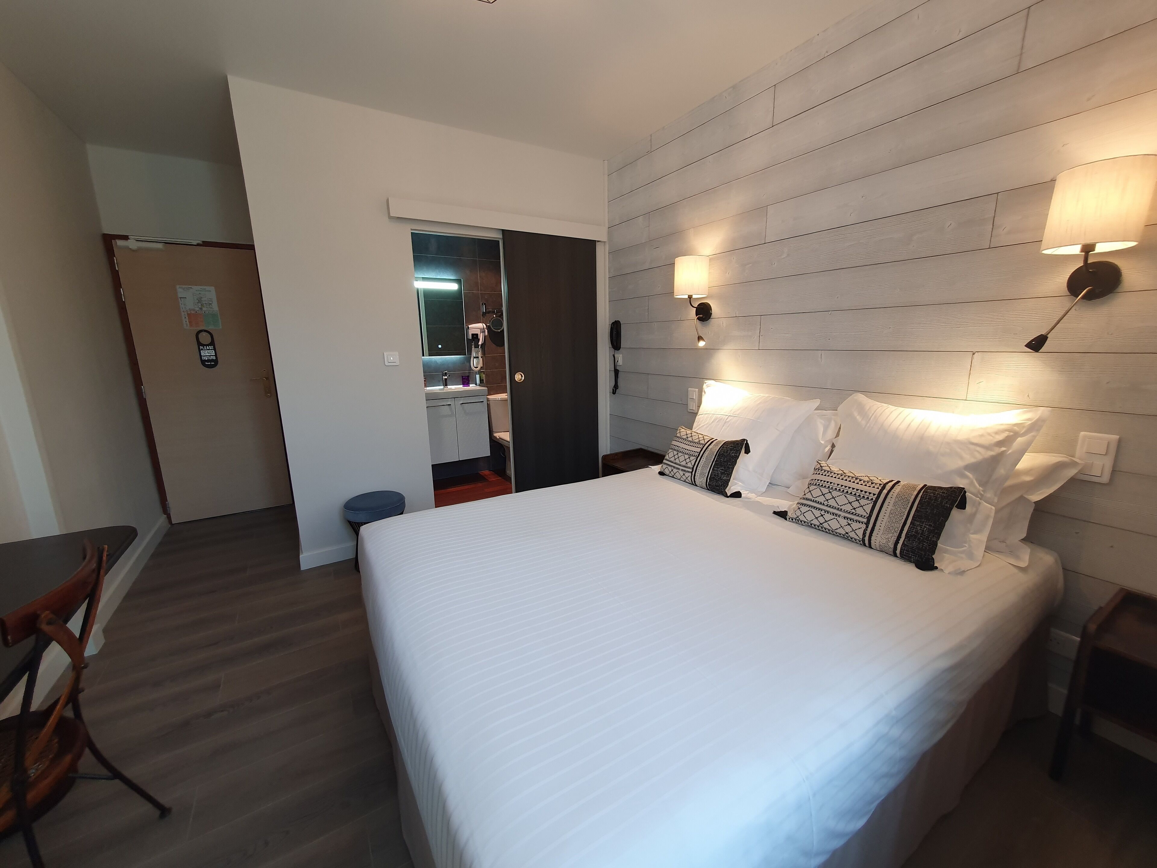 Quarto de hotel com uma cama grande, cabeceira de madeira, duas luminárias de mesa, uma mesinha e uma cadeira.