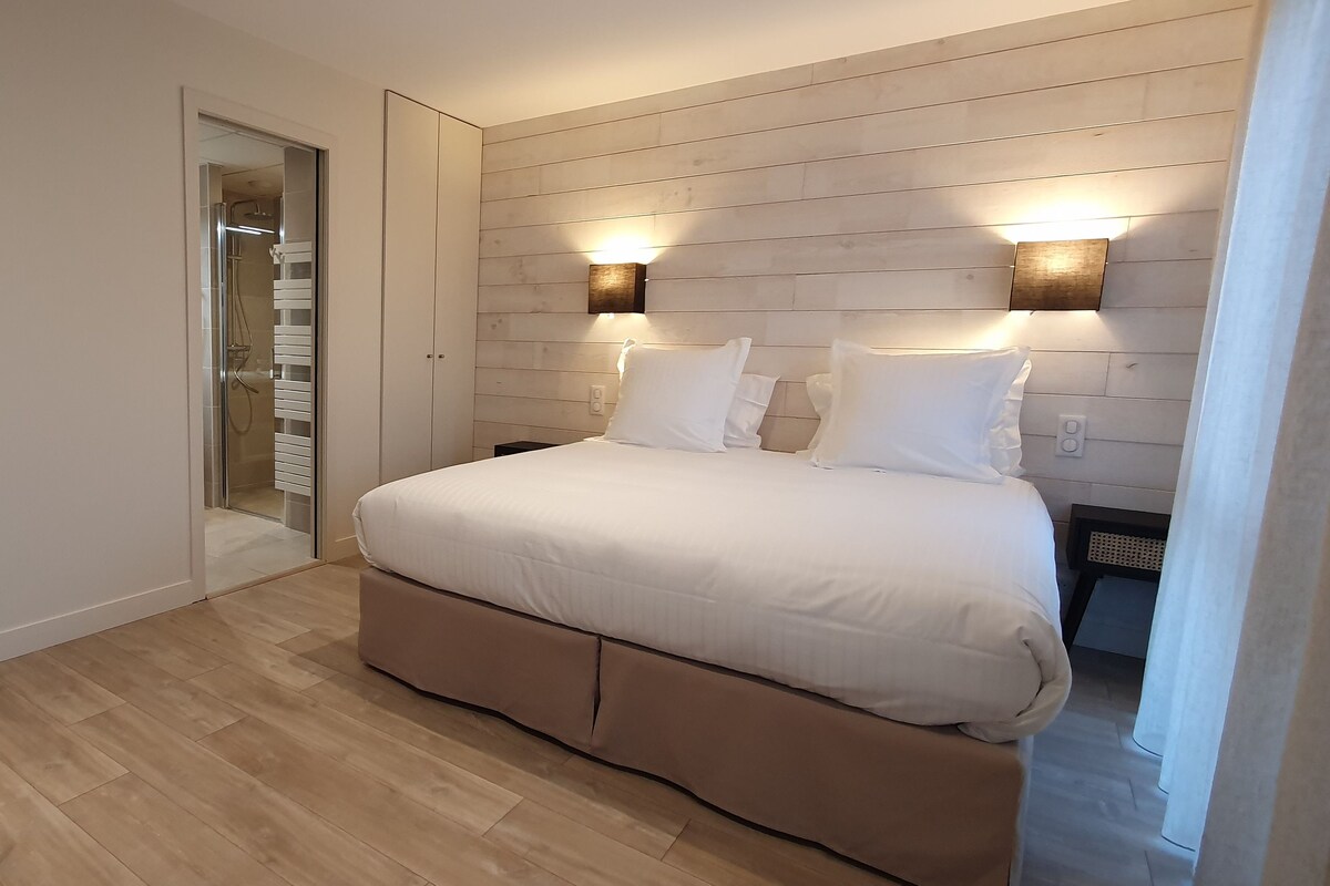 Een slaapkamer met een groot bed, houten vloeren, een glazen deur naar een badkamer en twee wandlampen.