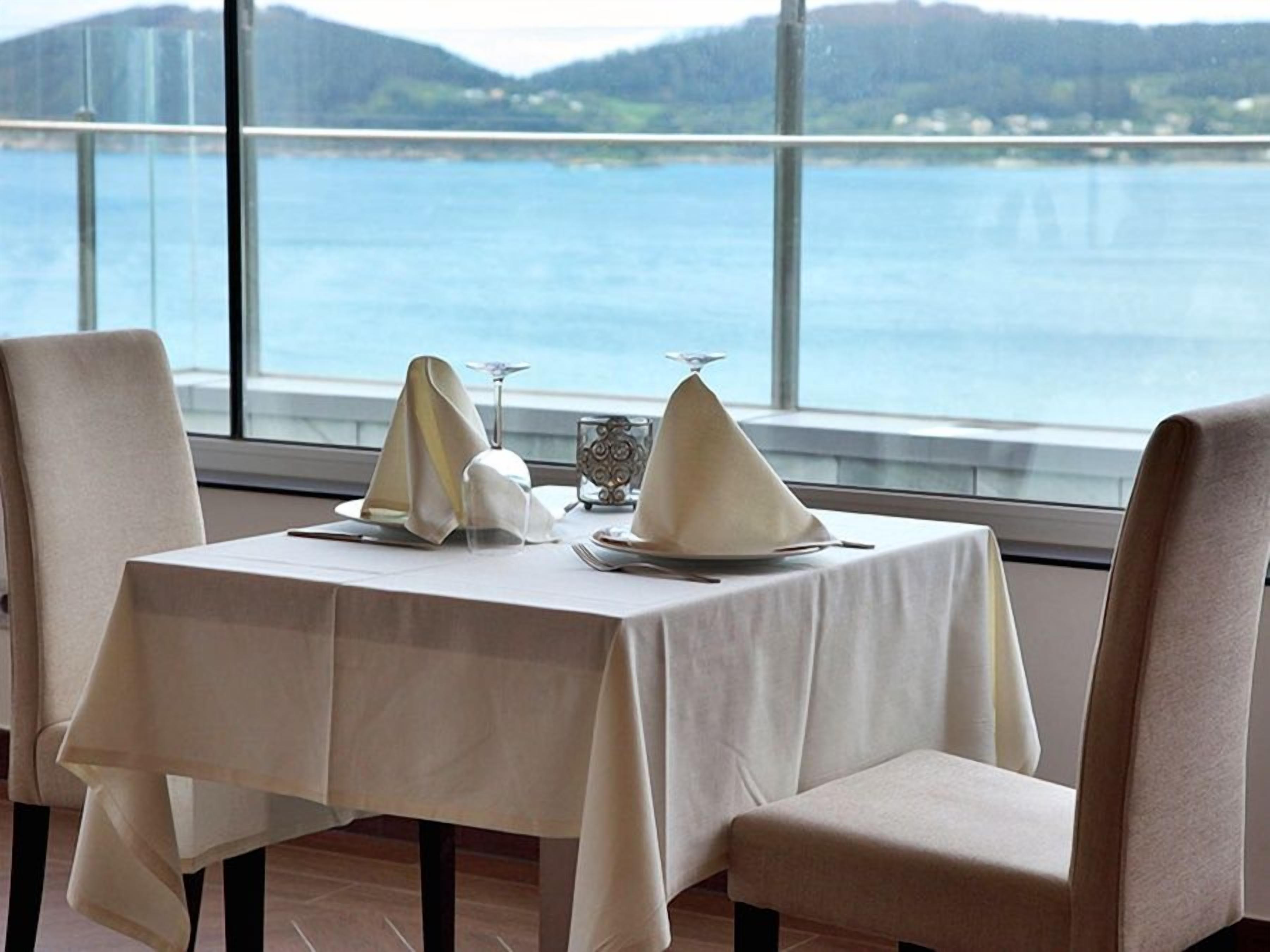 A dining table set with white tablecloths and plates, positioned near a window overlooking a body of water.