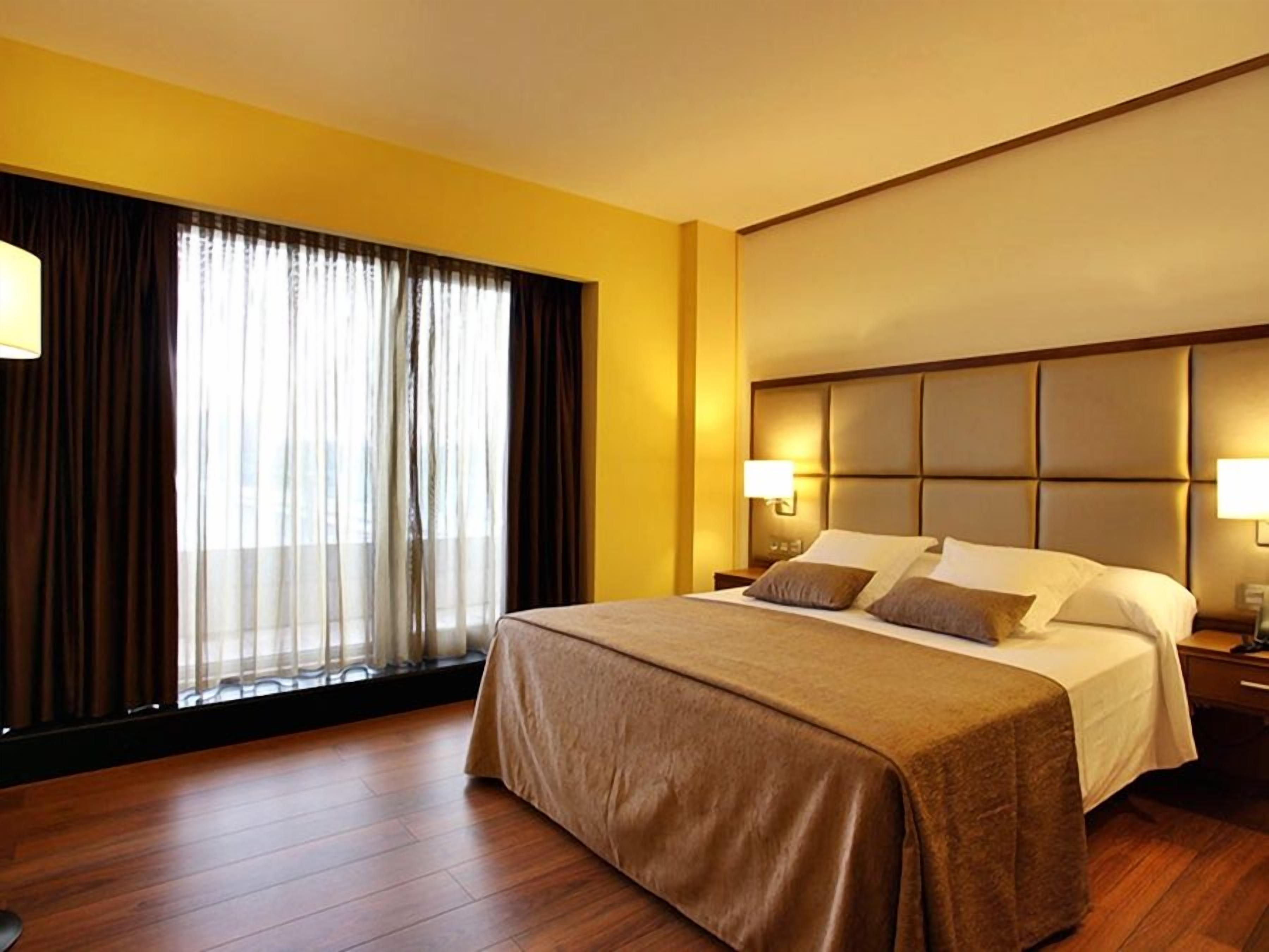 A hotel room with a large bed, a headboard with a padded design, a nightstand with a lamp, and a window with curtains.