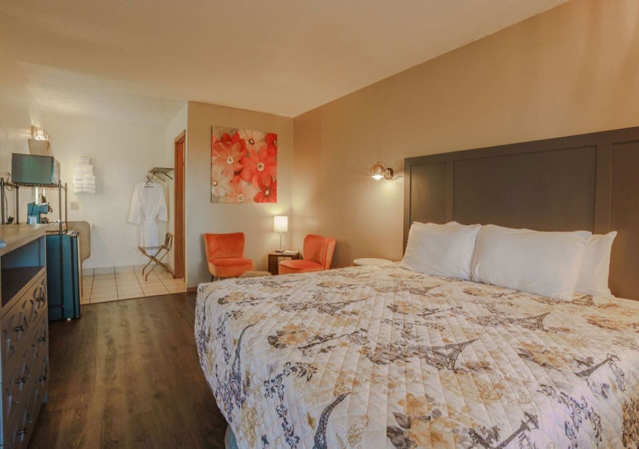 A hotel room with a large bed, a desk with a chair, a small table with a lamp, and a wall-mounted TV.