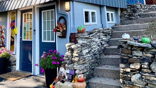 A two-story house with a blue exterior, a front porch with a welcome mat, and a stone wall at the entrance. - Shady Acre Inn and Suites (Branson West)
