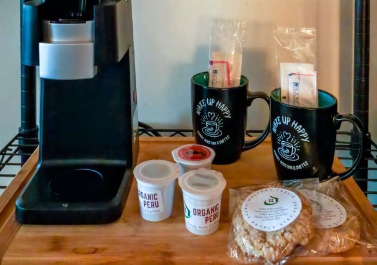 A coffee maker, two mugs, two containers of organic coffee, and two packages of cookies on a wooden tray.