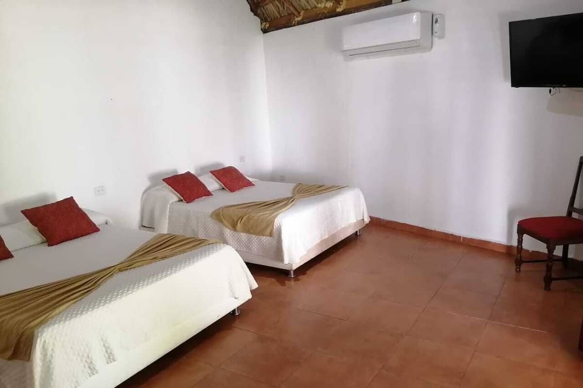 A room with two beds, each with a red pillow and a beige blanket. There is a TV mounted on the wall and a chair with a red cushion.