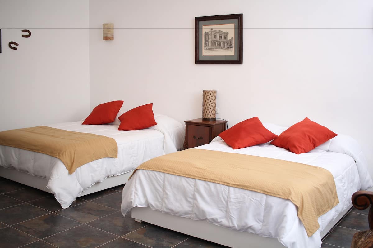 Two single beds with white bedding and red pillows, a wooden nightstand with a lamp, and a framed picture on the wall.