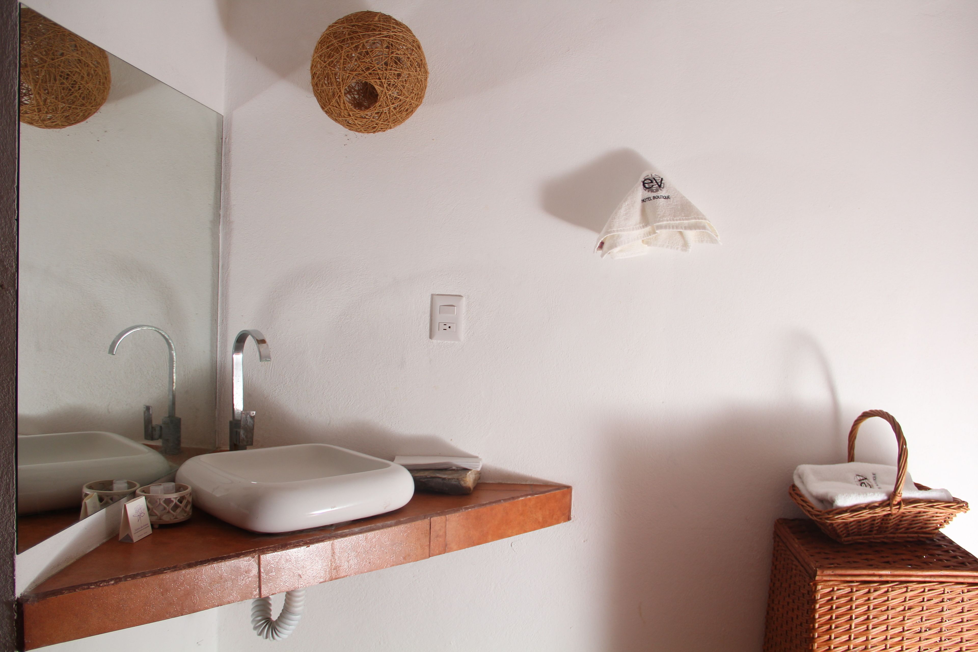 A bathroom with a white sink, mirror, and a wooden vanity. There is a wicker basket on the right and a woven light fixture on the wall.