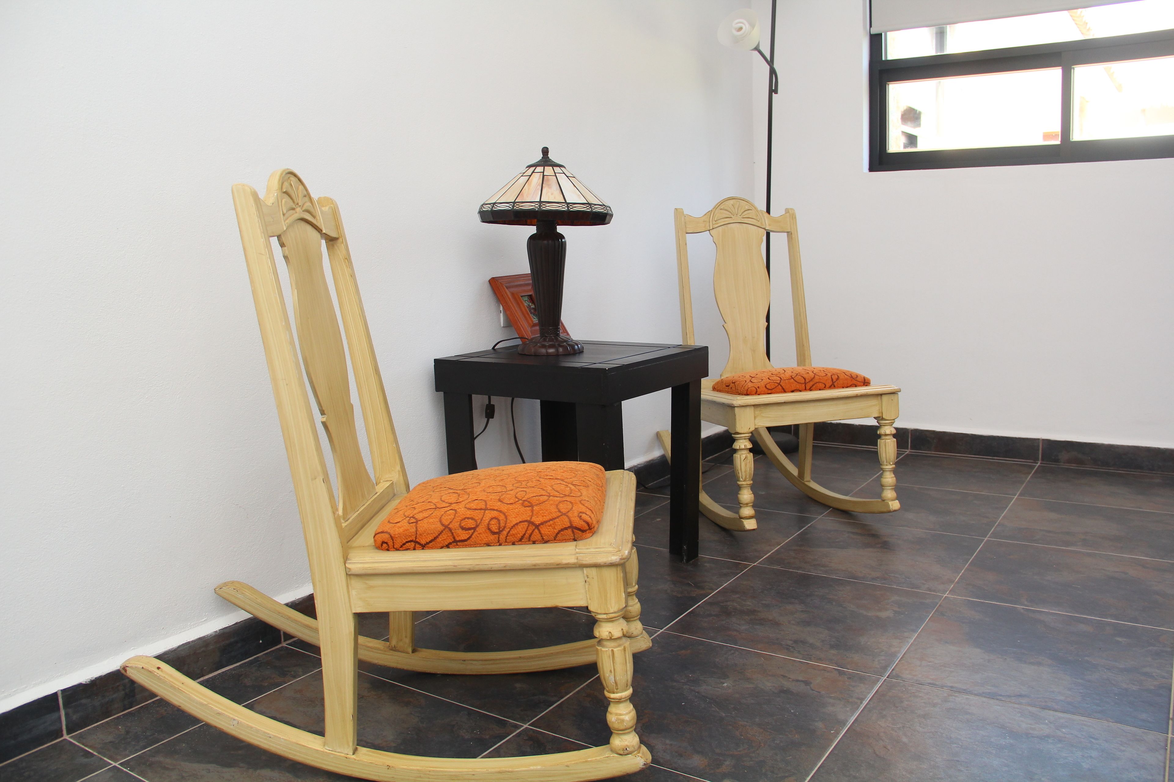 Two rocking chairs with orange cushions, a small table with a lamp, and a window with a view of the outside.