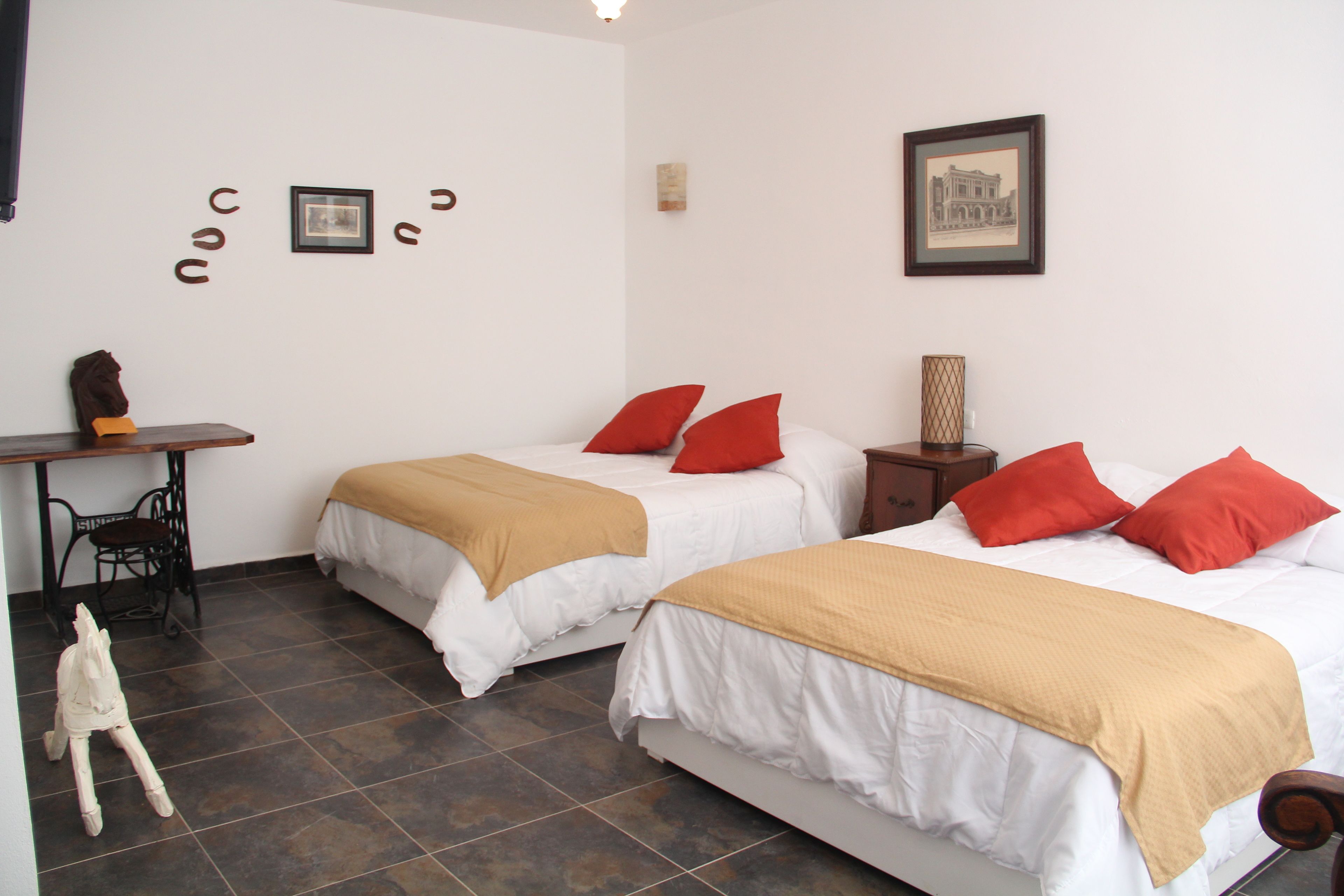 A hotel room with two beds, each with white bedding and red pillows, a desk with a chair, and a small statue on the floor.