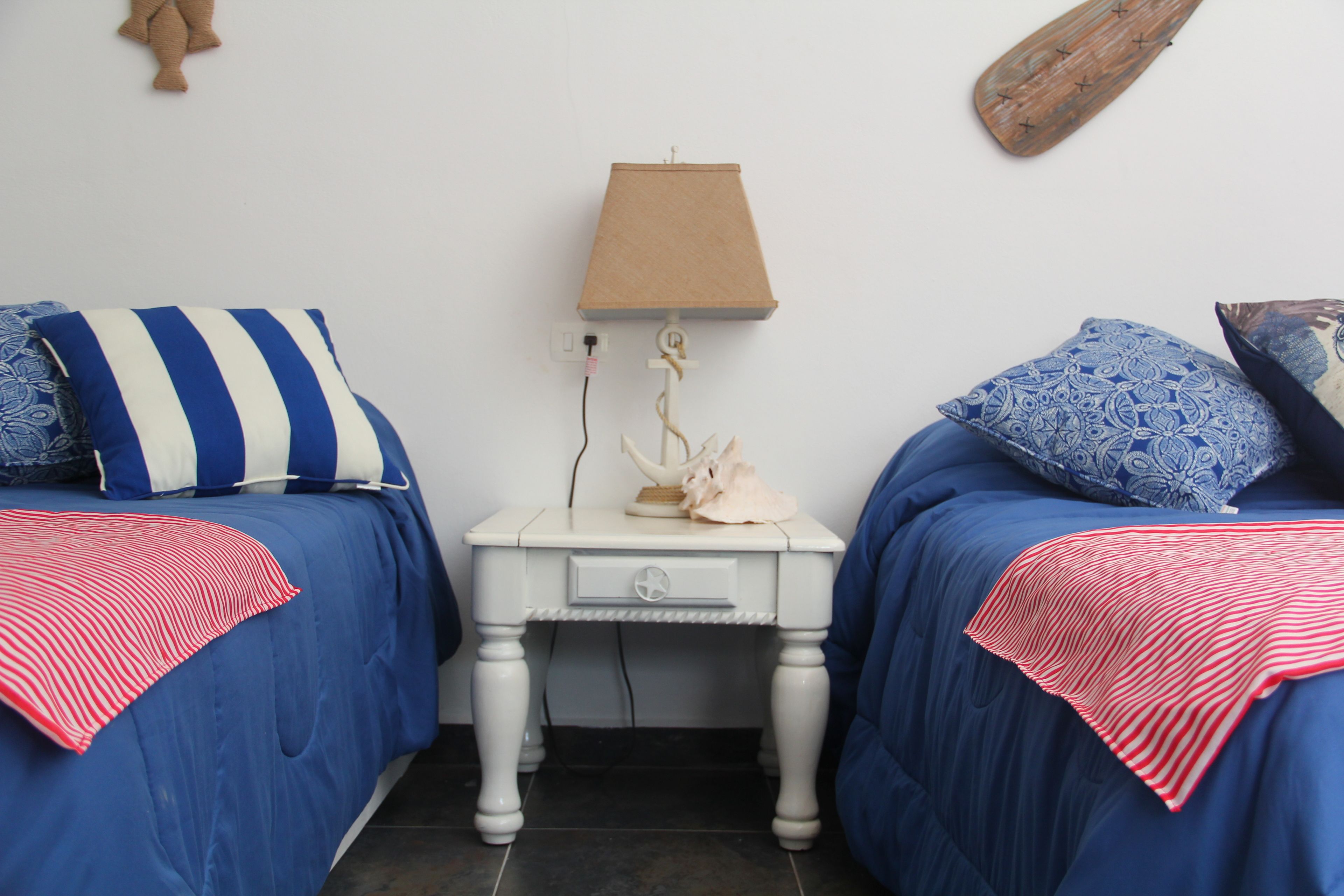 Un dormitorio con dos camas, cada una con una colcha azul y una almohada a rayas rojas y blancas. Hay una mesita de noche blanca con una lámpara y un objeto decorativo entre las camas.