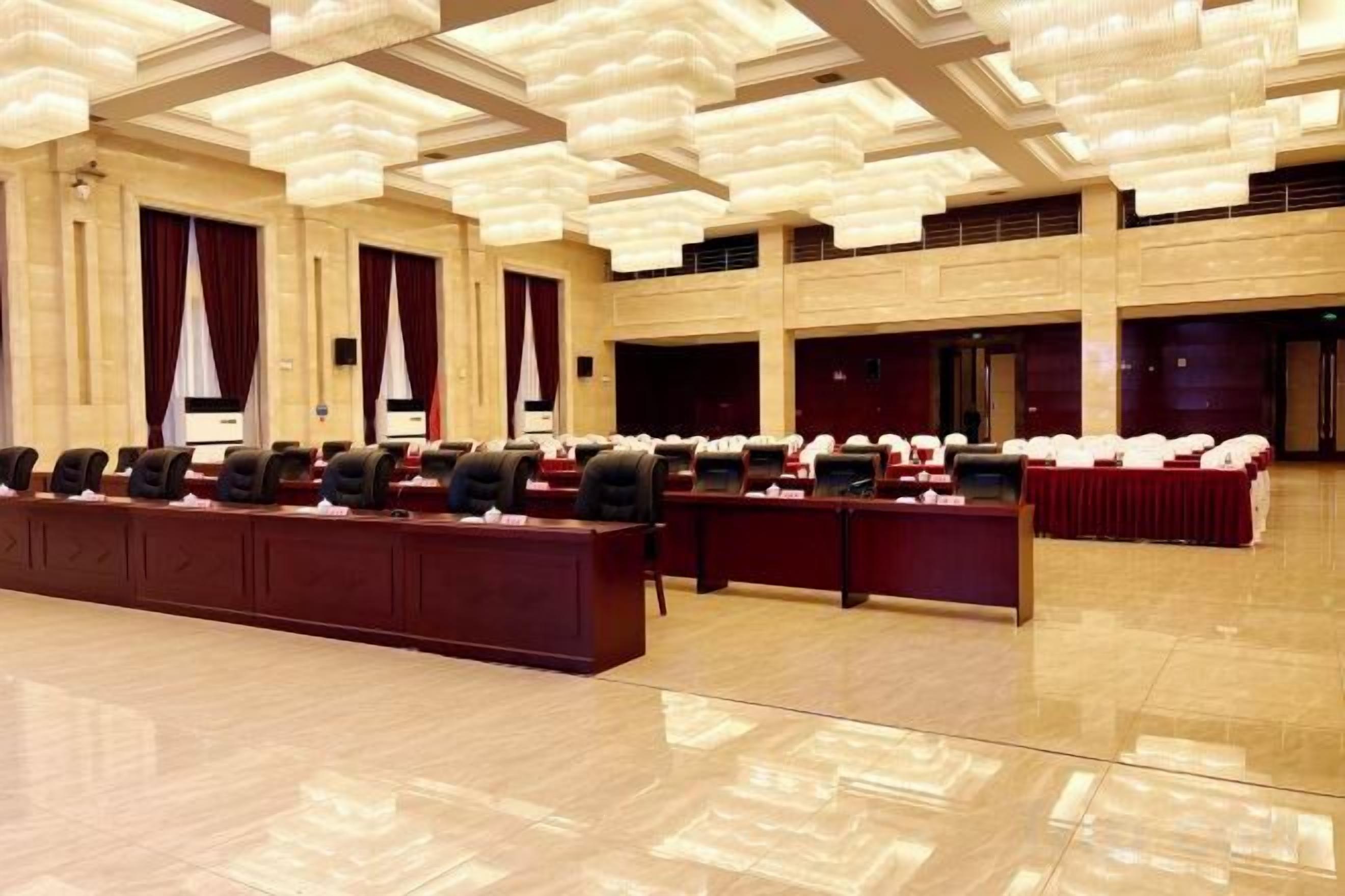 A conference room with multiple tables and chairs, a polished wooden floor, and a high ceiling with hanging lights.