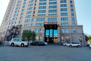 Un edificio de varios pisos con una entrada destacada, varios autos estacionados y un cielo despejado.