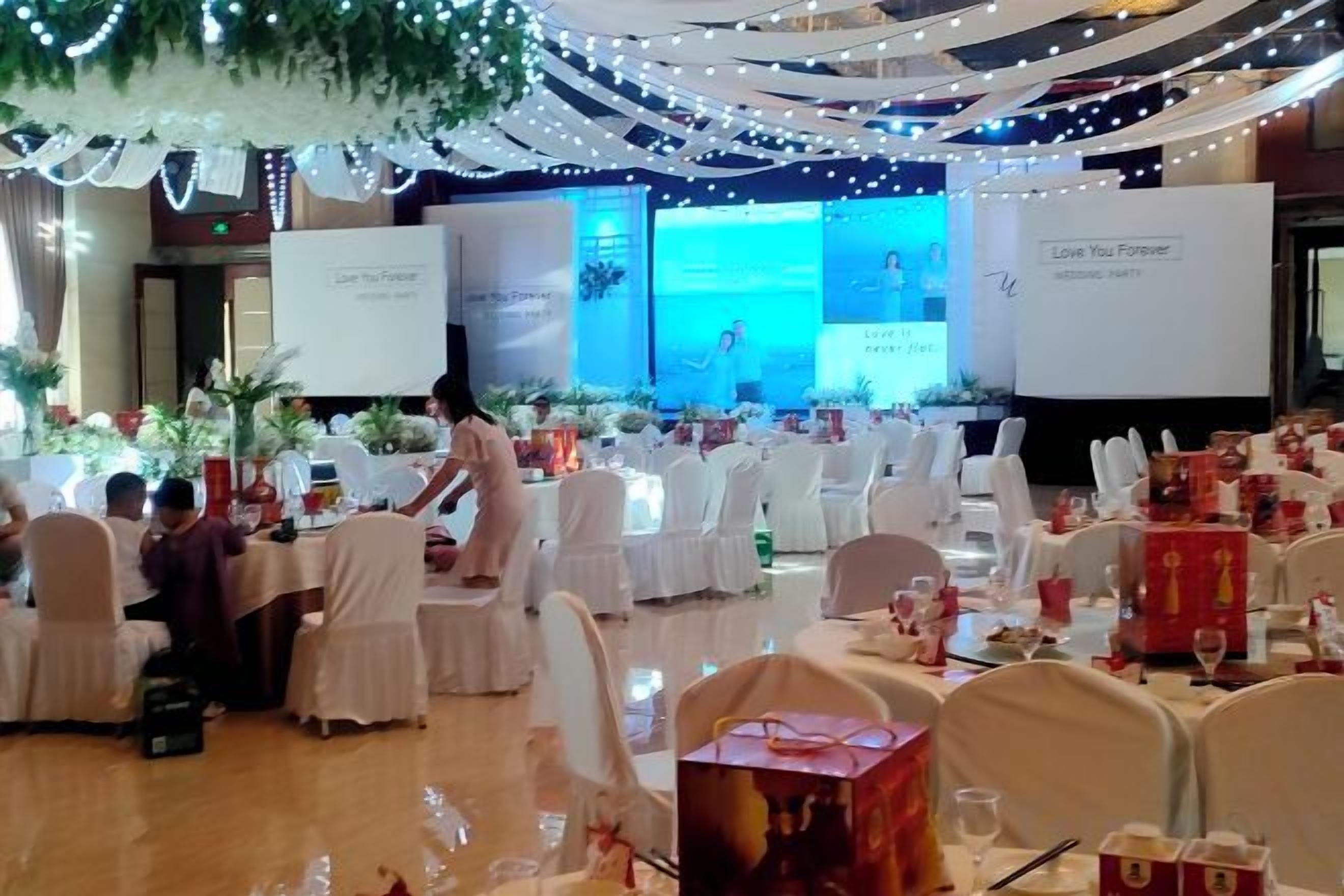 A banquet hall with round tables, white chairs, and a stage with a presentation screen.