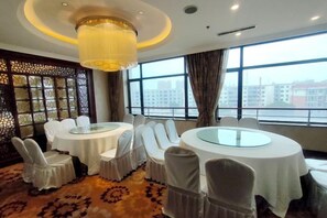 Habitación de hotel con dos mesas redondas, sillas blancas, una araña y grandes ventanales con vista a edificios.