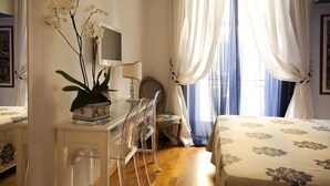 A bedroom with a bed, a desk with a chair, a potted orchid, a television, and a window with curtains.