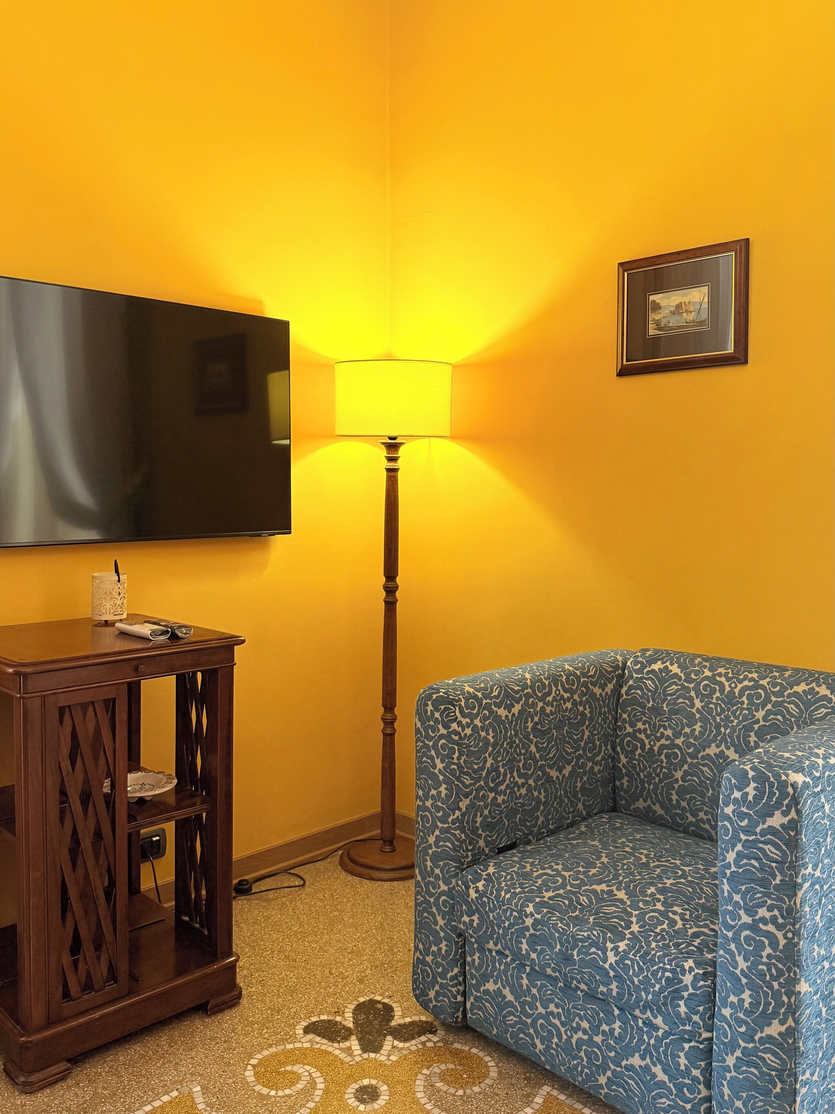 Ein Zimmer mit einem an der Wand befestigten Fernseher, einer stehenden Stehlampe, einem gemusterten Sofa und einem kleinen Holztisch mit einem dekorativen Gegenstand.