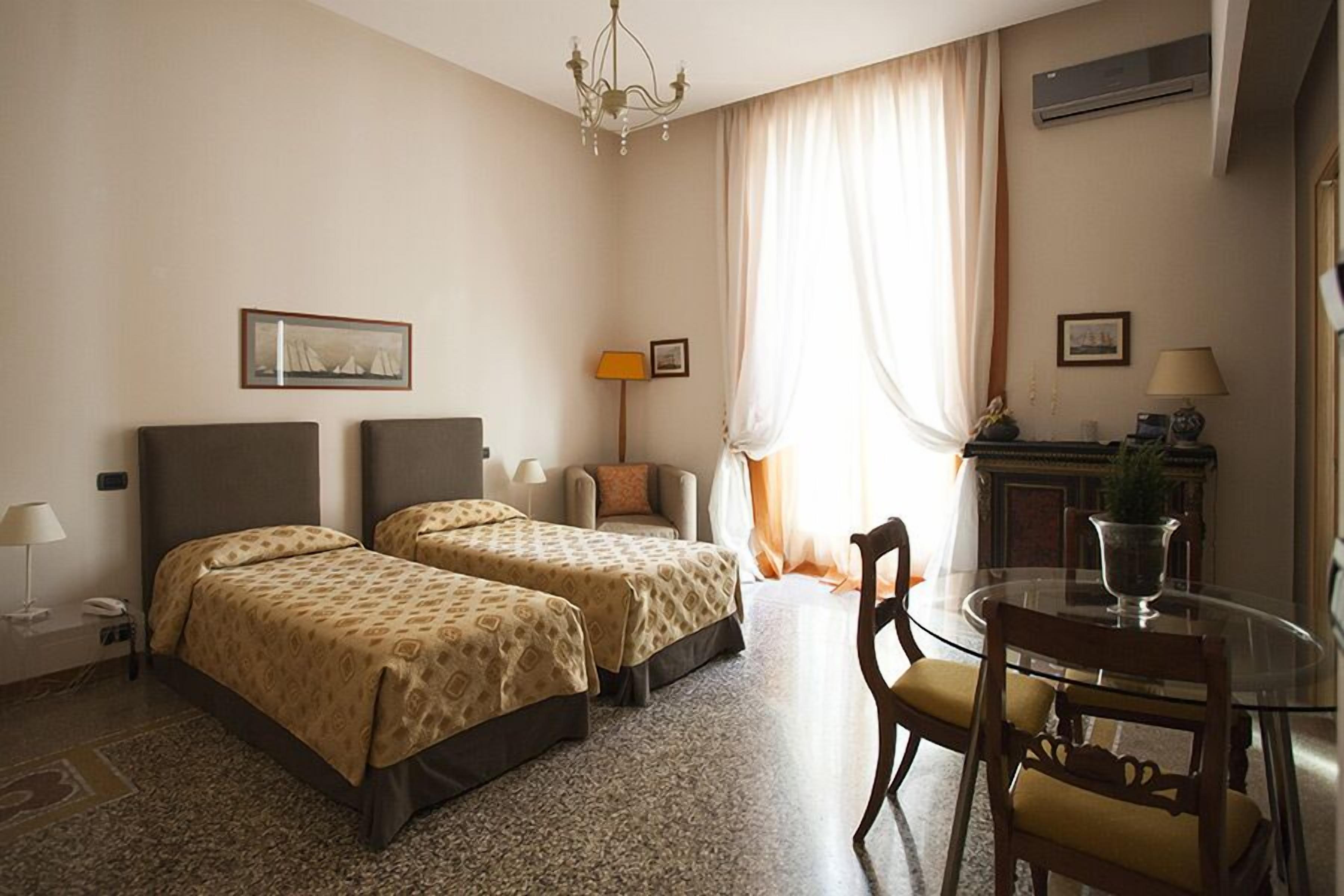 A hotel room with two beds, a dining table with chairs, a chandelier, and a fireplace.