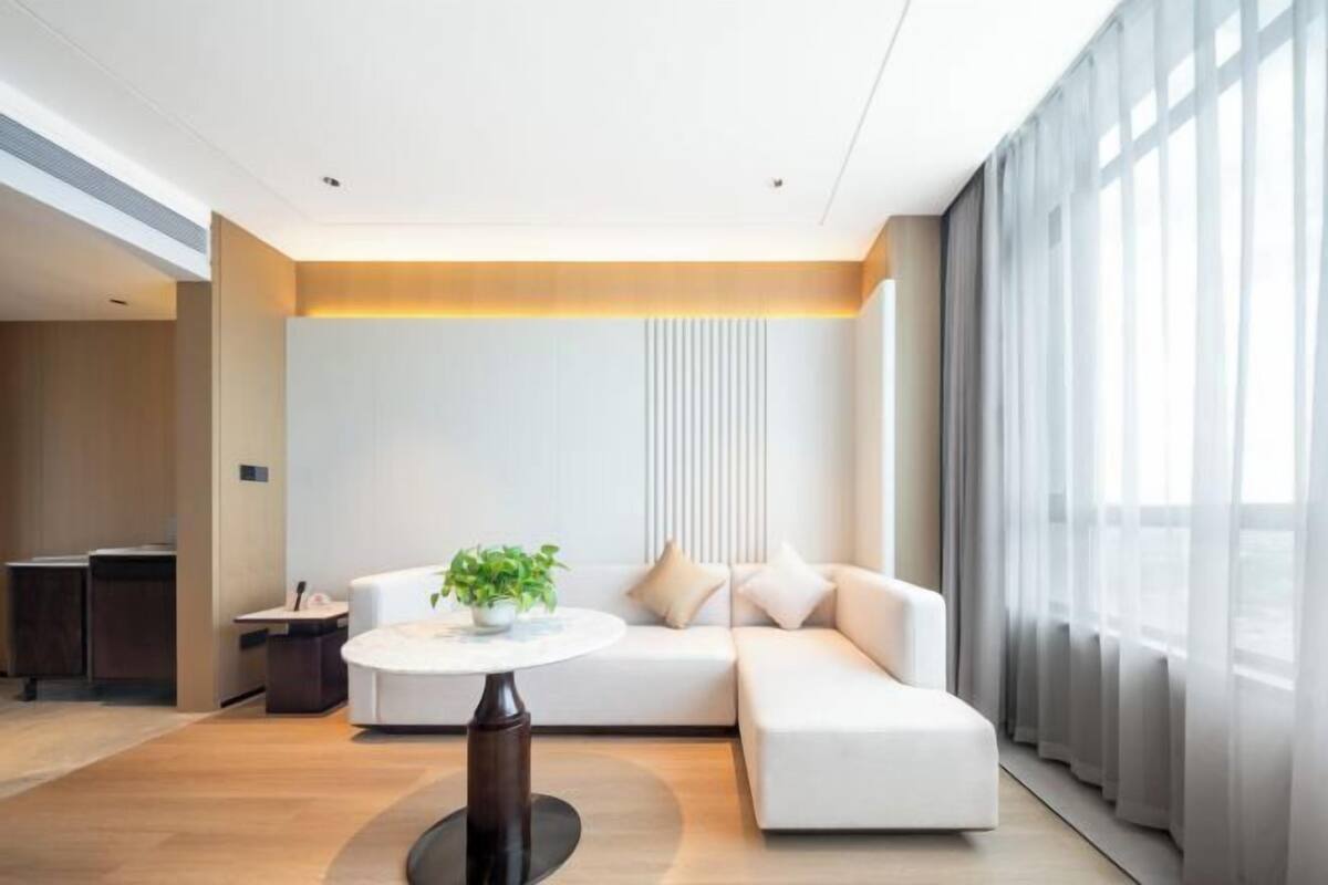 A modern living room with a white sofa, a round table with a plant, and a large window with sheer curtains.