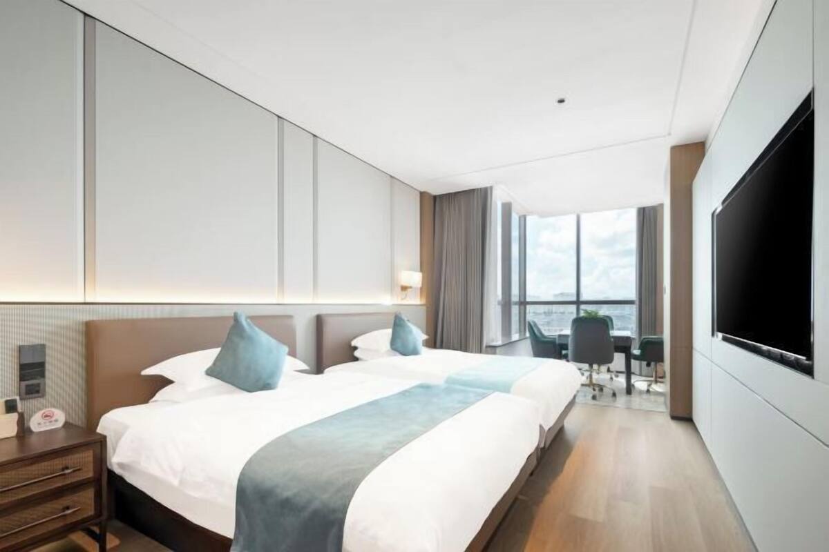 A hotel room with a large bed, a desk, a chair, a TV, and a view of the city.