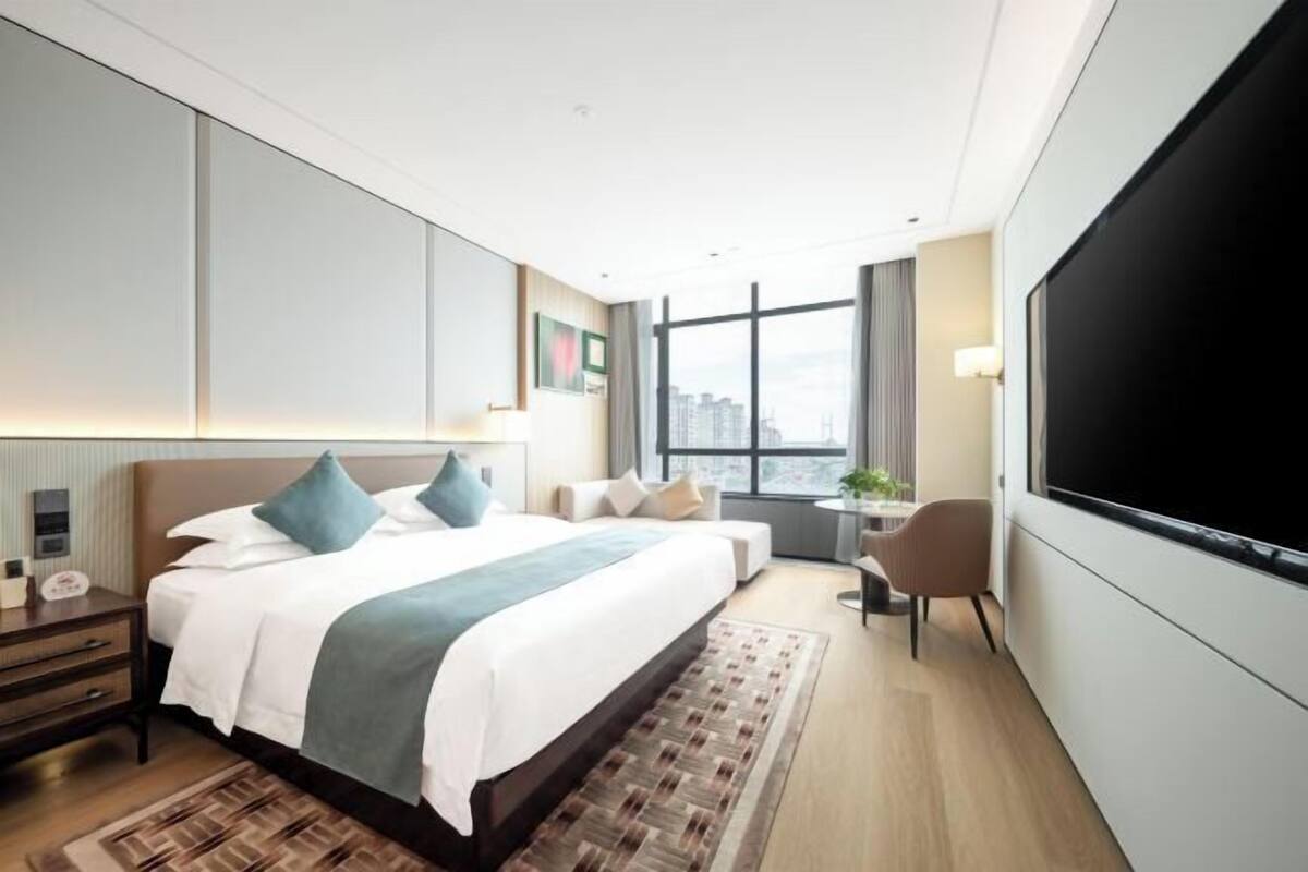 A modern hotel room with a large bed, a TV, a desk, and a view of the city.