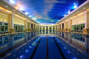 Una amplia piscina interior con un carril central, rodeada de zonas de descanso y un techo con un diseño de iluminación azul.
